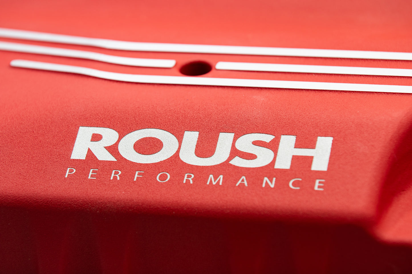 Close-up of a red surface with the white text "ROUSH Performance" and decorative white lines above it, hinting at the power of an 810-horsepower ROUSH Mustang Supercharger.