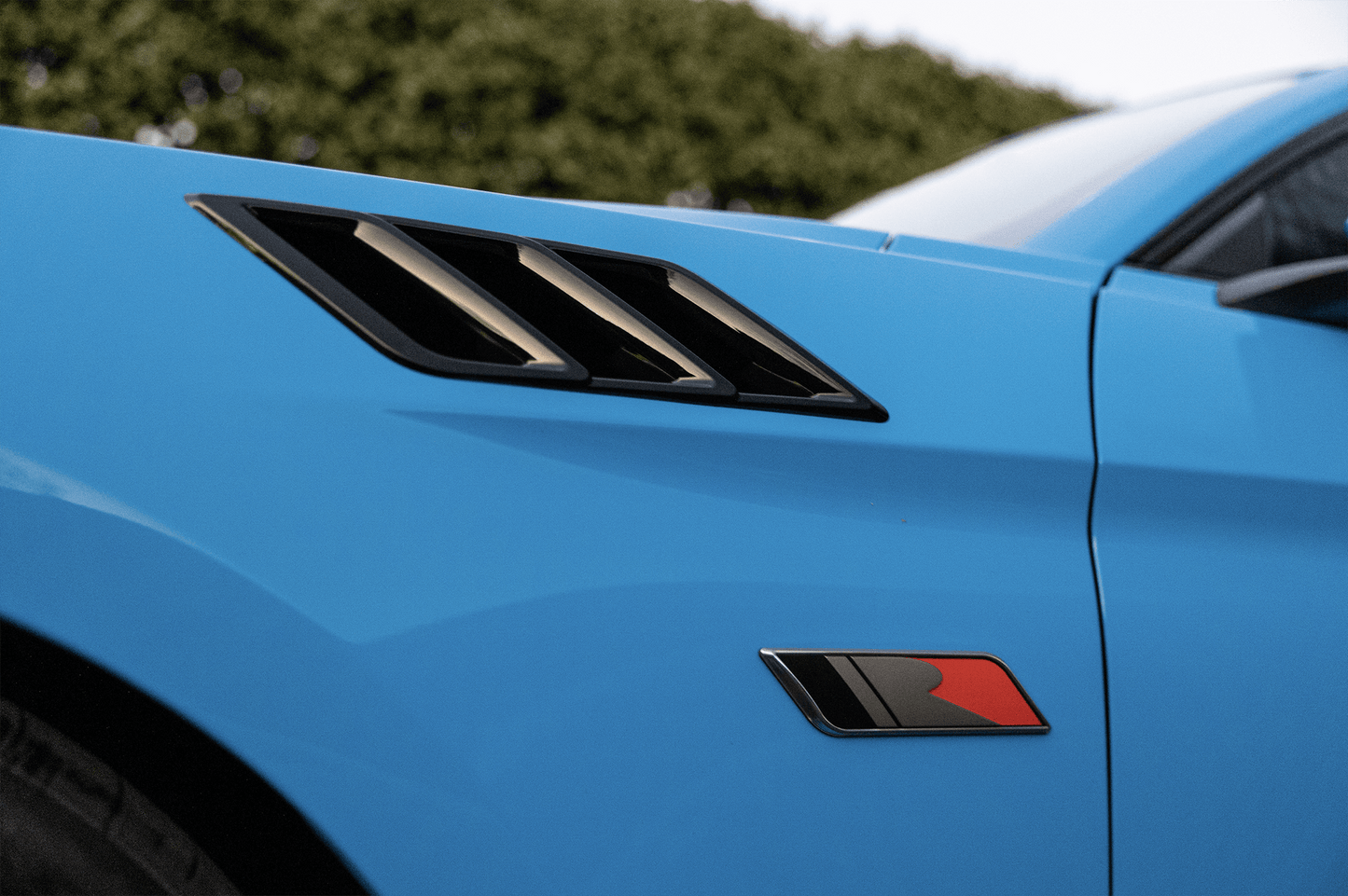 Close-up of a blue Roush Mustang showcasing high-performance design. Its sleek black fender vent heat extractors complement a small emblem below, featuring a striking red and black motif.