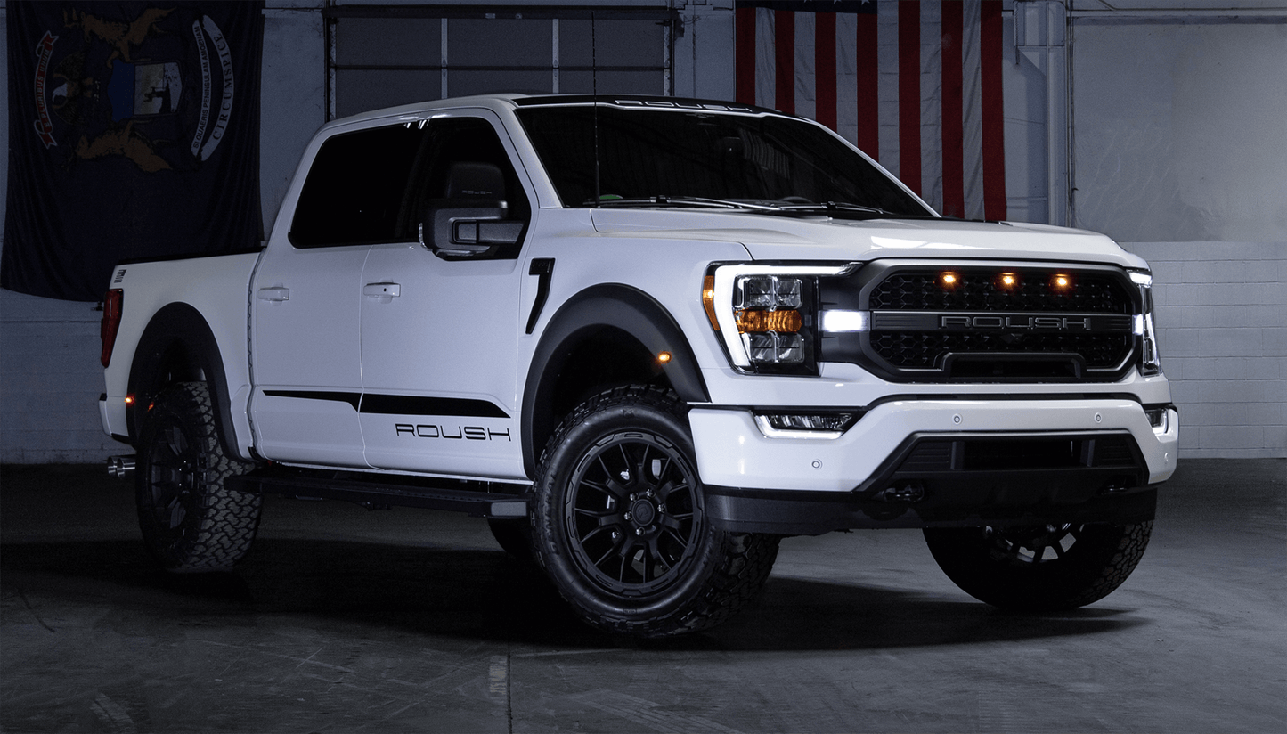 A white pickup truck with a "Roush" logo is parked indoors, showcasing matte black rims and front grille. It features 20-inch Roush F-150 wheels. An American flag and state flag elegantly adorn the background.