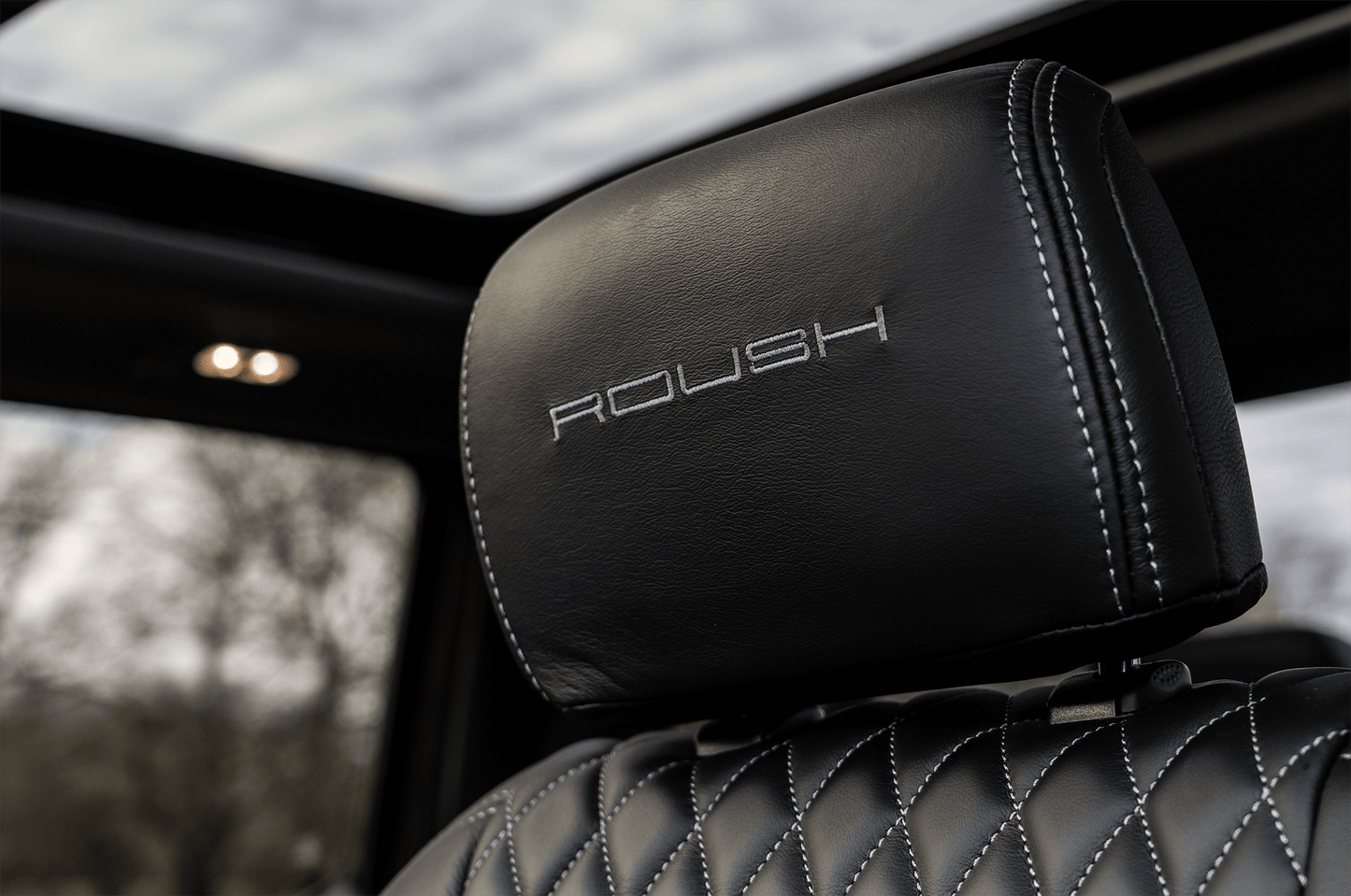 Close-up of a black leather car seat headrest with the word "ROUSH" embroidered in white stitching, with a view of the sunroof and cloudy sky visible above. The distinctive ROUSH High-Flow Front Grille of the 2022 ROUSH F-150 hints at the power and performance ready to be unleashed.