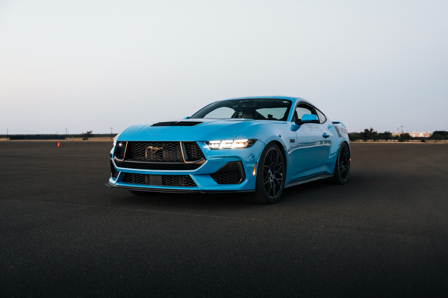 A blue sports car, equipped with aggressive drop and Roush Mustang Lowering Springs, is parked on a wide, empty asphalt surface under a clear sky.