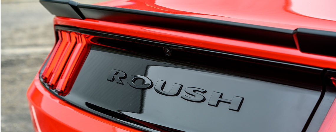 Rear view close-up of a red Mustang featuring a black spoiler and "ROUSH" prominently displayed on the trunk, emphasizing its performance pedigree.
