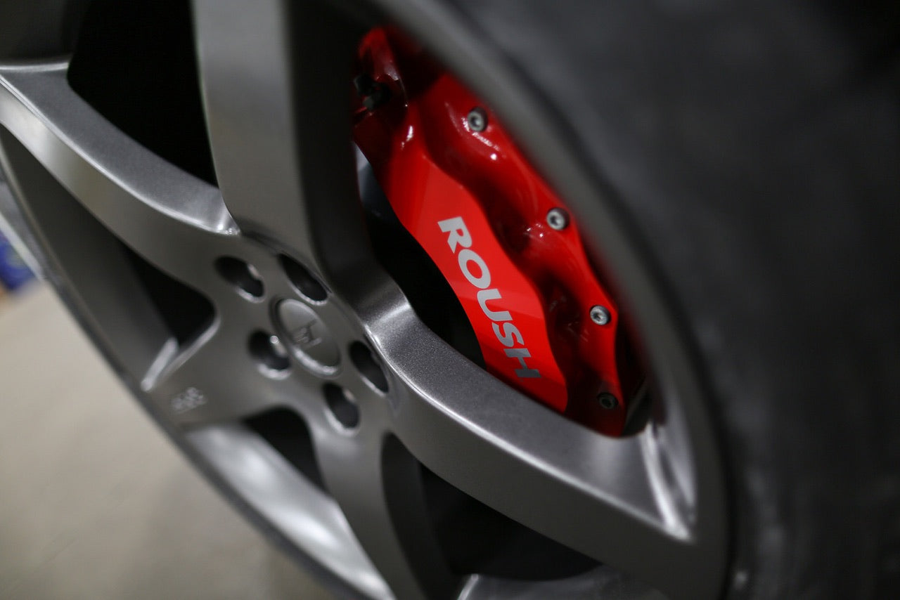 Close-up of a car wheel featuring a metallic gray rim and a red brake caliper with the word "ROUSH" visible, capturing the essence of the ROUSH Stage 2 Mustang's V8 power.