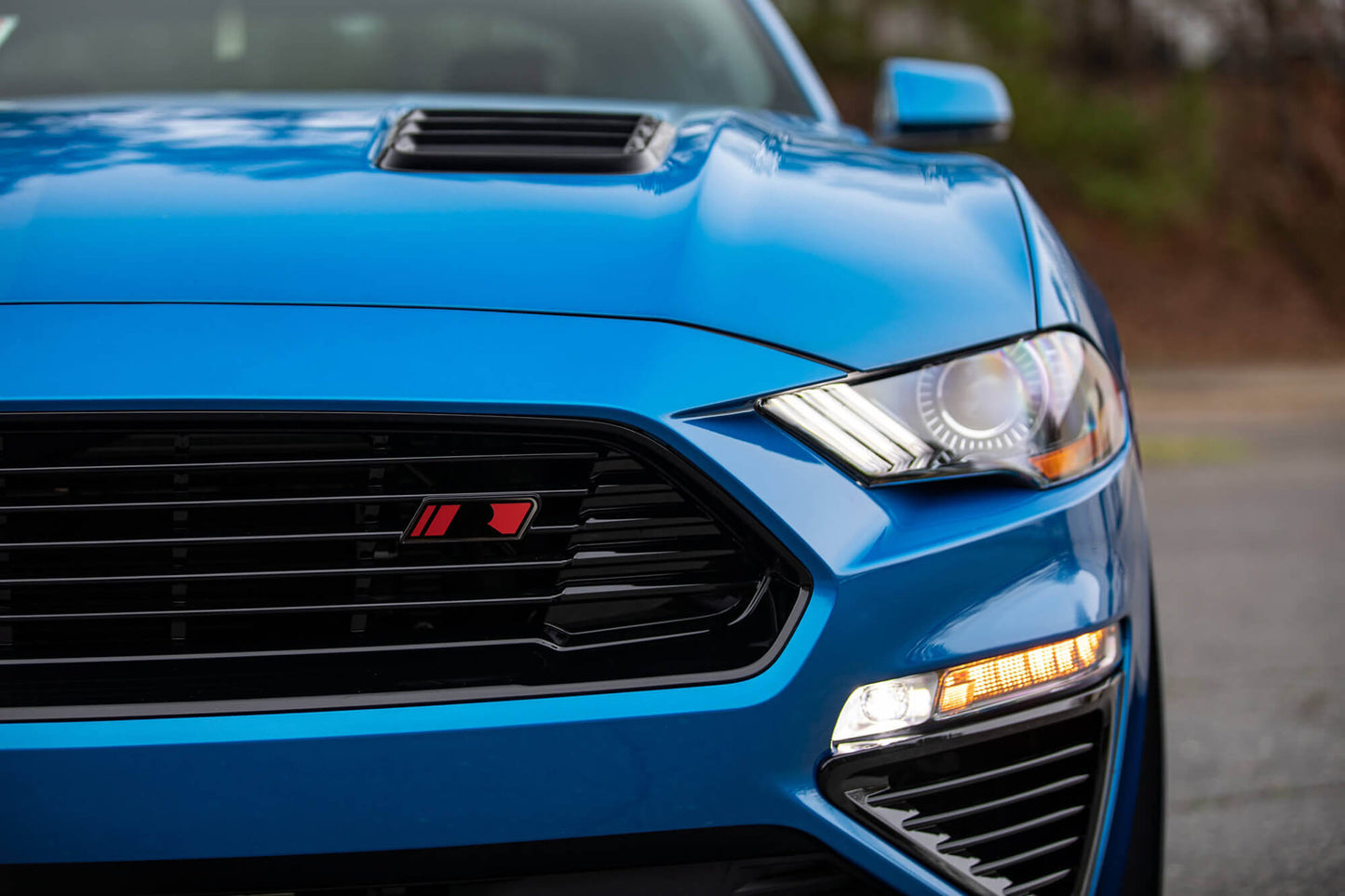 Close-up of the front end of a blue sports car showing the Mustang front grille, headlights, and part of the hood with a vent in a high gloss black finish.