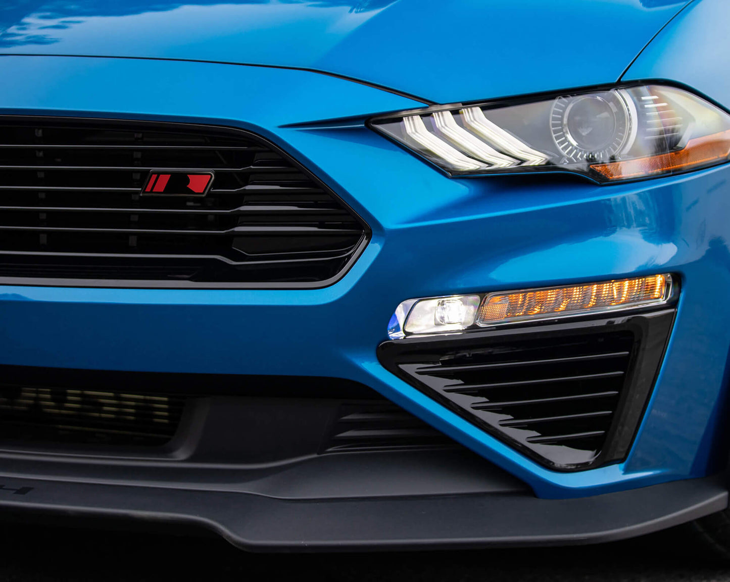 Close-up shot of the front end of a blue sports car, featuring the Roush Mustang Front Grille and highlighting the high gloss black finish on the headlight, grill, and bumper design.