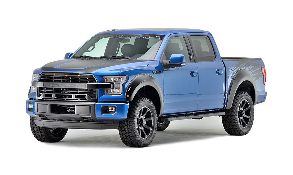 A ROUSH F-150, boasting black accents and large tires, is parked against a white background. Enhanced with the ROUSH Performance Series Suspension System, this blue pickup truck promises superior handling and style.