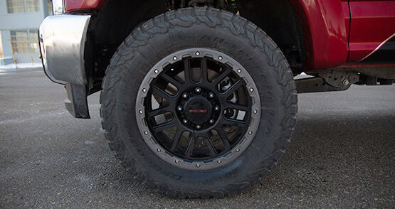 A close-up of a large black off-road tire and rim on a red ROUSH Super Duty vehicle, parked on a paved surface, showcasing its robust performance and impressive towing capabilities.