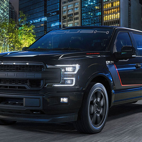 A black pickup truck with a bold grille and red accents, featuring a ROUSH Performance exhaust system, roars down a city street at night, surrounded by tall buildings.