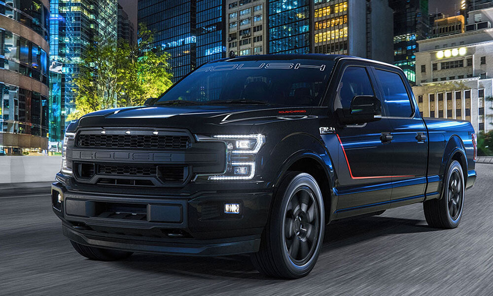A black pickup truck with a bold grille and red accents, featuring a ROUSH Performance exhaust system, roars down a city street at night, surrounded by tall buildings.