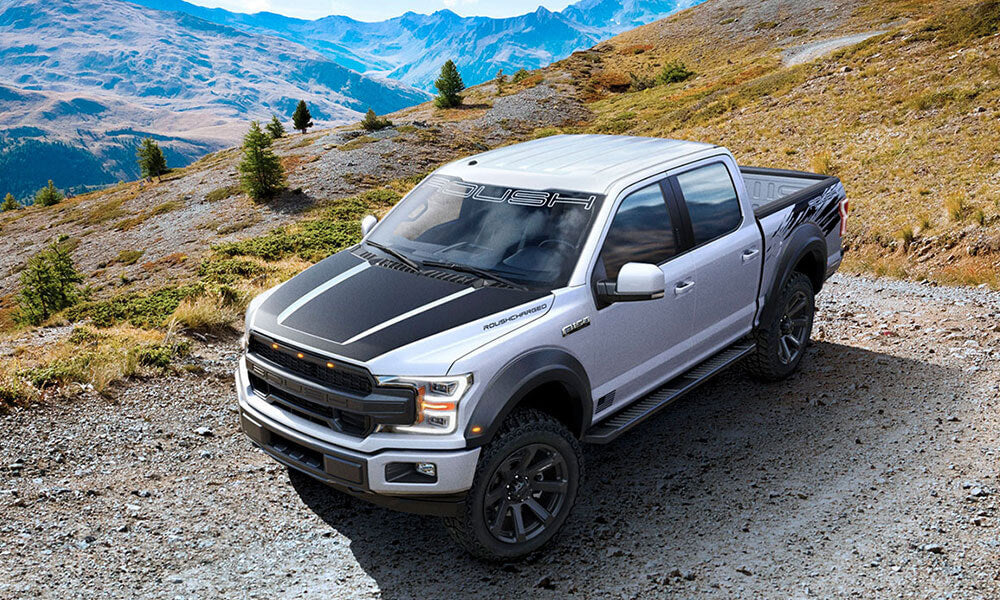 A silver pickup truck with ROUSH Performance black stripes sits proudly on a rocky path in a mountainous landscape, showcasing its impressive off-road capability.