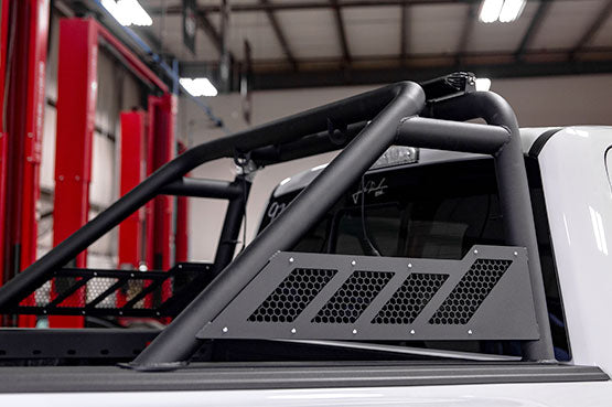 
                  
                    Close-up of a truck with a black roll bar and mesh panel, showcasing its ROUSH Raptor build, parked indoors under fluorescent lights.
                  
                