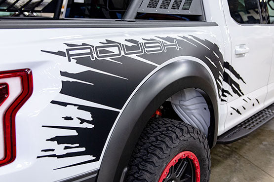 
                  
                    Close-up of a white ROUSH Raptor truck featuring bold black graphics and the word "Roush" on the side, with visible rear wheel and fender equipped with off-road wheels.
                  
                