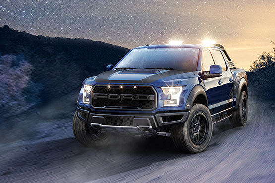
                  
                    A blue ROUSH Raptor pickup truck with headlights on rumbles along a dirt road at dusk, its off-road wheels expertly handling the rugged terrain against a mountainous backdrop.
                  
                