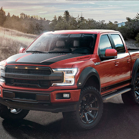 A ROUSH F-150 SC, with its performance exhaust system, is parked on a rural road, surrounded by dry grass and trees under a cloudy sky. The red and black pickup truck stands out amidst the rugged landscape.
