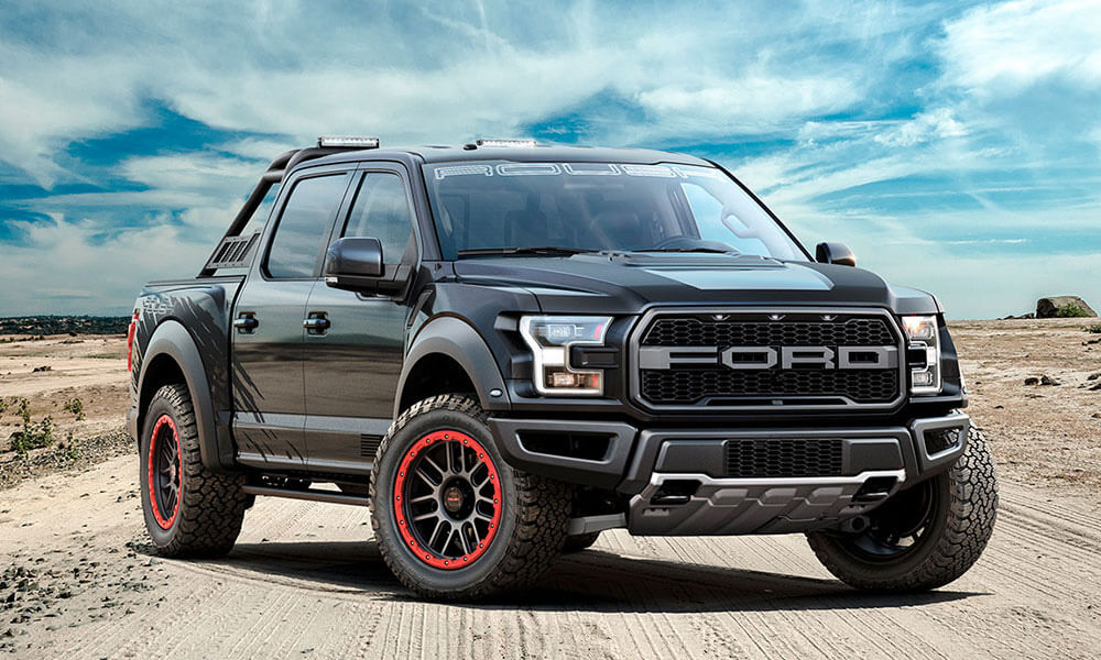 A black Ford ROUSH Raptor pickup truck with red-accented off-road wheels is parked on a sandy terrain under a blue sky.