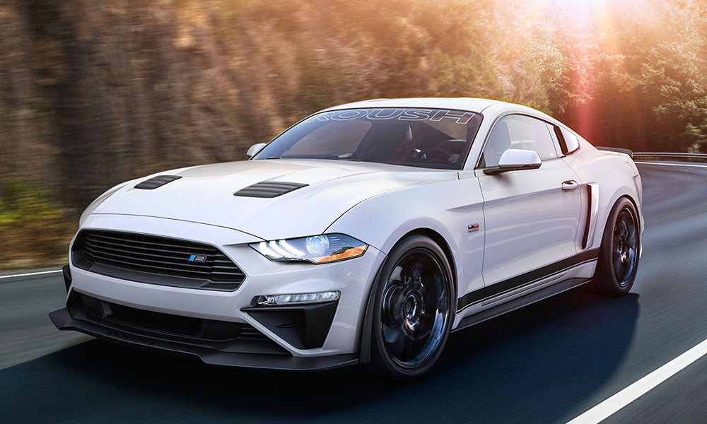 
                  
                    A white Mustang with a "ROUSH" decal and a roaring 5.0L V8 engine glides effortlessly on a forested road in daylight.
                  
                