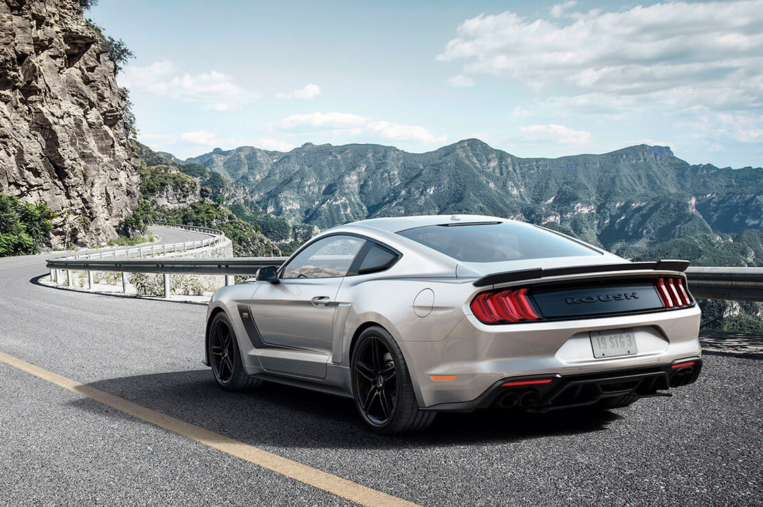 
                  
                    A silver sports car with a ROUSH Stage 3 supercharger roars down a winding mountain road, its active exhaust system echoing through the green hills, all under a partly cloudy sky.
                  
                