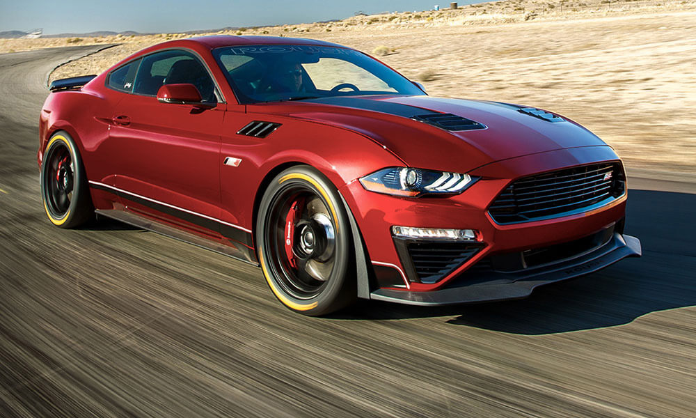 
                  
                    A red ROUSH Mustang with 775HP supercharger roars down the desert road, captured from the front side. This Jack Roush Edition sports car showcases black accents and sleek design elements, embodying power and style.
                  
                