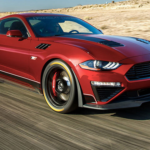 A red ROUSH Mustang with 775HP supercharger roars down the desert road, captured from the front side. This Jack Roush Edition sports car showcases black accents and sleek design elements, embodying power and style.