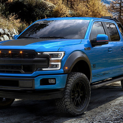 The 2020 ROUSH F-150 SC, with its supercharged power and robust off-road suspension, navigates effortlessly along a gravel road flanked by trees and rocks in a picturesque mountainous area.