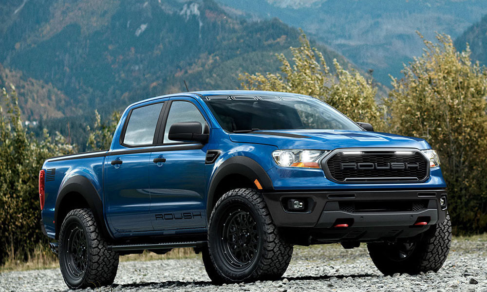 A blue ROUSH Ranger pickup truck, showcasing its off-road prowess and performance suspension system, is parked on a gravel surface with mountains and trees in the background.