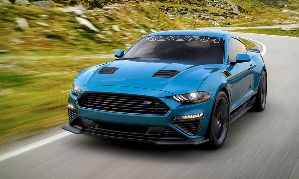 A blue Mustang with "ROUSH" emblazoned on the windshield speeds along a winding road, slicing through the grassy, rocky landscape with unmatched performance.