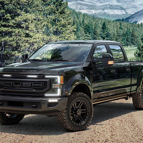 A black pickup truck equipped with all-terrain tires on a dirt road is surrounded by a dense forest and majestic mountains in the background.