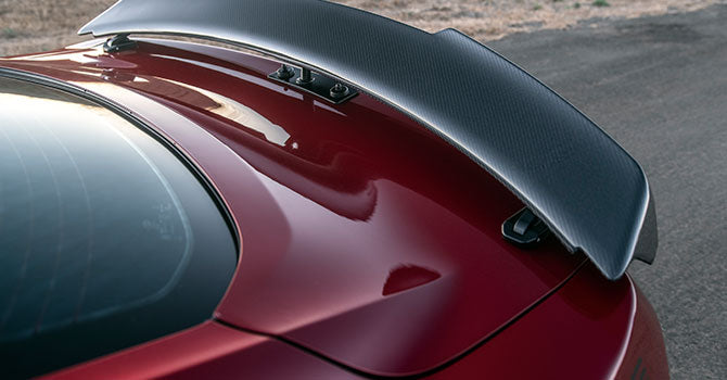 
                  
                    Rear view of a red car with a large black spoiler on a road, showcasing its ROUSH Stage 3 enhancements and supercharger for an exhilarating drive.
                  
                