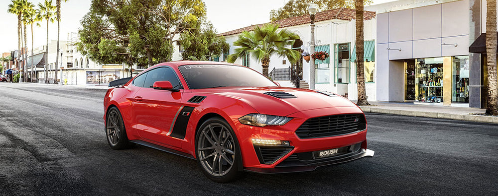 
                  
                    A red sports car equipped with a ROUSH Stage 3 supercharger is parked on a palm-lined street, with quaint shops and lush trees in the background.
                  
                