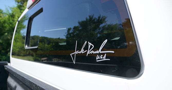 
                  
                    The rear window of a white vehicle features a white signature decal reading "Jack Roush USA," hinting at the off-road prowess of the ROUSH Ranger. Lush trees are visible in the background, enhancing this rugged scene.
                  
                