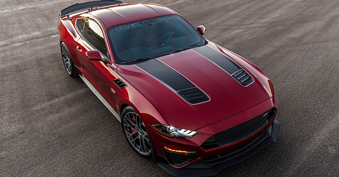 
                  
                    The ROUSH Mustang, in a striking Jack Roush Edition, showcases its red body with black racing stripes on the hood. With its 775HP supercharger, this powerful sports car is parked on a paved surface and viewed from an elevated angle.
                  
                