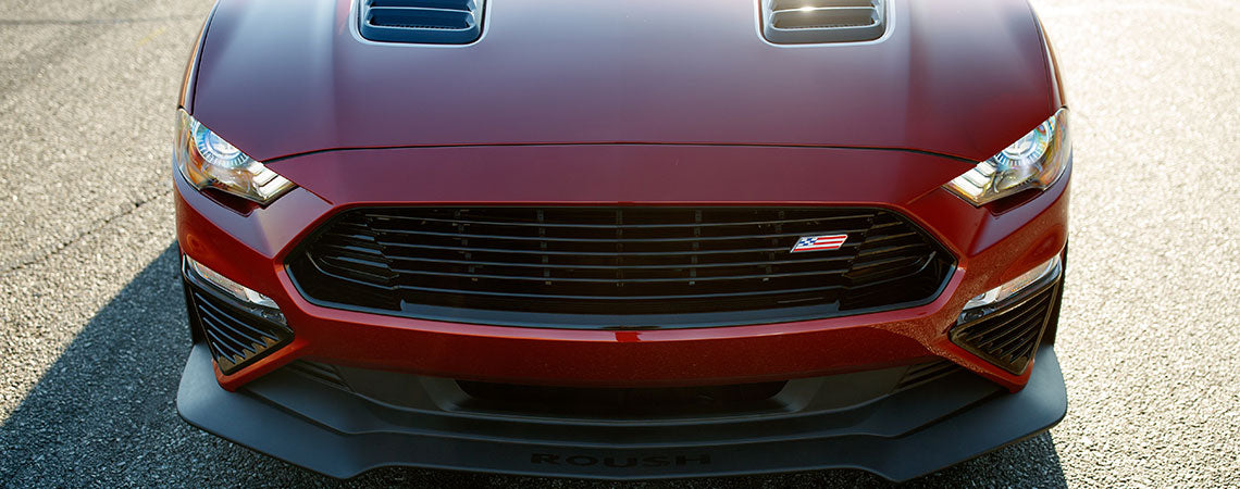 
                  
                    Front view of a red ROUSH Mustang sports car with a black grille, dual hood vents, and a gleaming Roush emblem on a sunny day, showcasing its 775HP supercharger power.
                  
                