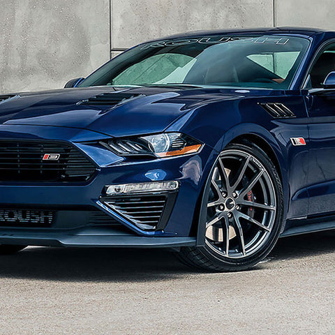 A dark blue 2021 ROUSH Stage 3 Mustang, boasting 750 horsepower, is parked on a concrete surface next to a light gray wall.