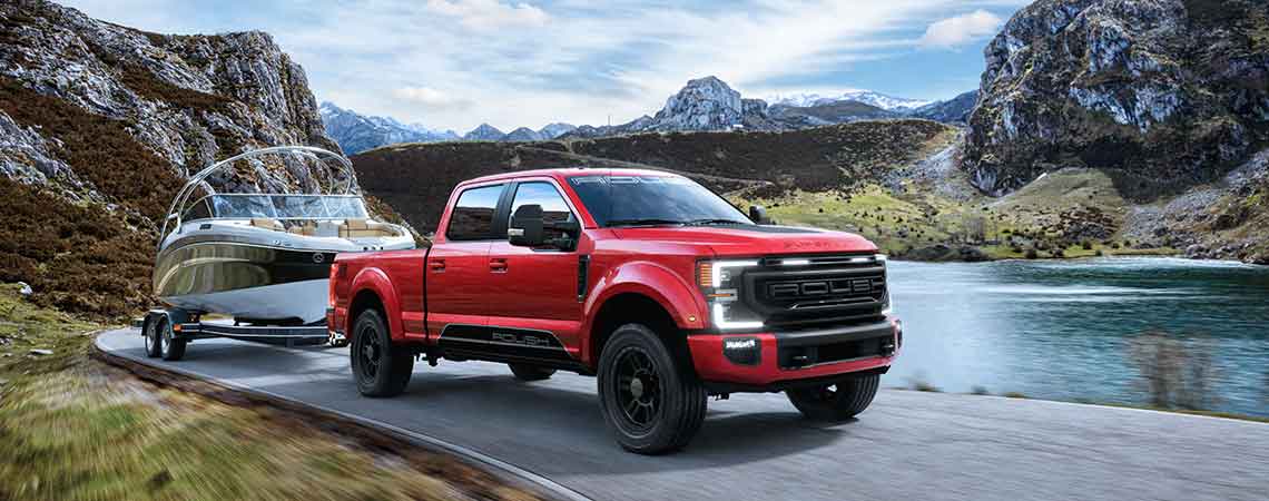 
                  
                    A red pickup truck with high-performance suspension and 20-inch ROUSH wheels tows a boat along a road beside a lake, framed by majestic mountains in the background.
                  
                