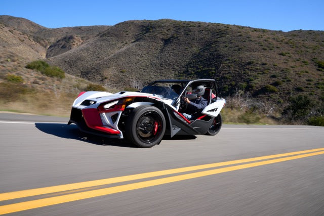 A three-wheeled, open-air Slingshot Roush Edition with red, black, and white colors drives on a paved road through a hilly landscape, showcasing its racetrack attitude on a sunny day.