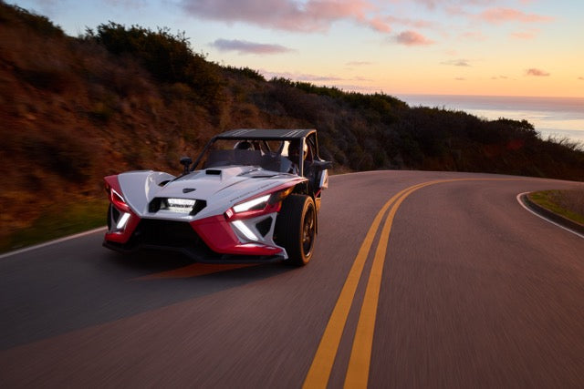 A Slingshot Roush Edition, the ultimate performance vehicle, drives on a winding road at sunset, with cliffs and the ocean in the background—its racetrack attitude unmistakable against the scenic vista.