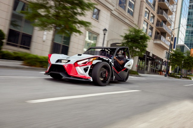 
                  
                    A three-wheeled, open-air Slingshot Roush Edition, boasting a red, white, and black design, is driven down a city street with modern buildings in the background, exuding racetrack attitude.
                  
                