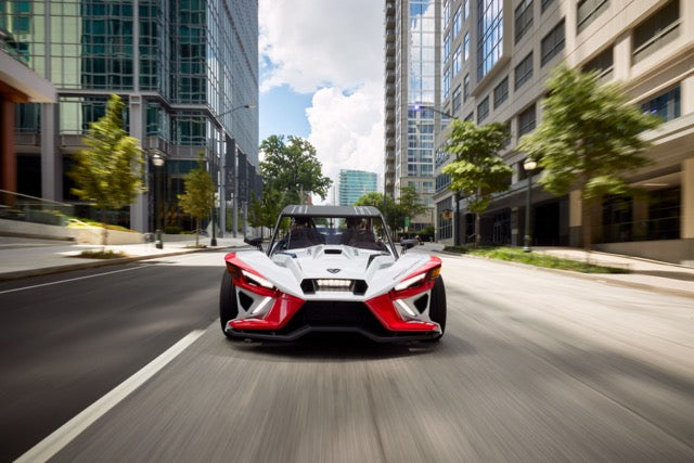 
                  
                    A three-wheeled, open-air Slingshot Roush Edition performance vehicle drives down a city street lined with modern high-rise buildings and trees.
                  
                