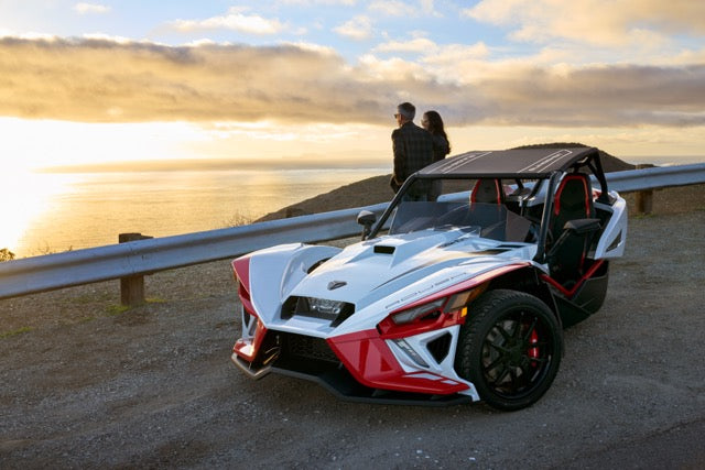 
                  
                    A three-wheeled Slingshot Roush Edition performance vehicle is parked by a roadside overlooking the ocean at sunset, with two people standing near the guardrail in the background.
                  
                