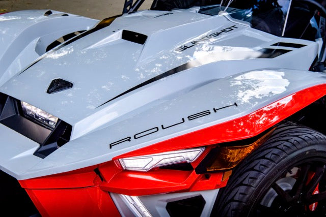 
                  
                    Close-up of the front end of a white and red Slingshot Roush Edition three-wheeled performance vehicle, highlighting its angular design details, bold branding, and unmistakable racetrack attitude.
                  
                