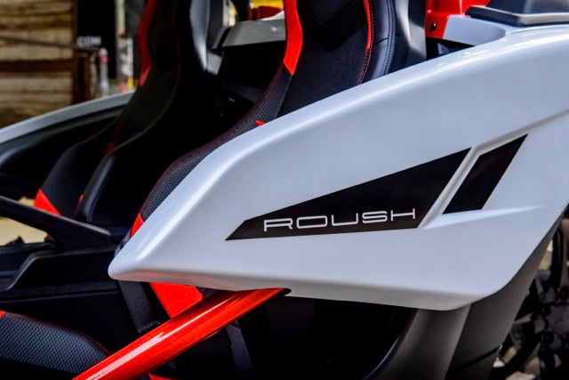 
                  
                    Close-up of a white Slingshot Roush Edition panel with the brand name "ROUSH" printed on it; part of a seat and red frame are visible in the background, emphasizing this performance vehicle’s racetrack attitude.
                  
                