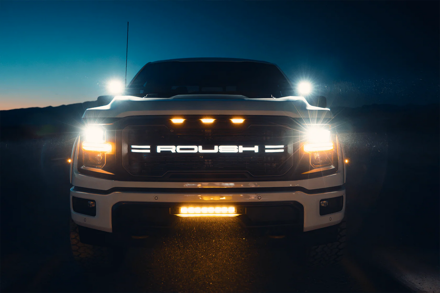 A 2024 Roush F-150 pickup with "ROUSH" illuminated on the grille, off-road wheels, headlights and roof lights on, photographed from the front at dusk.