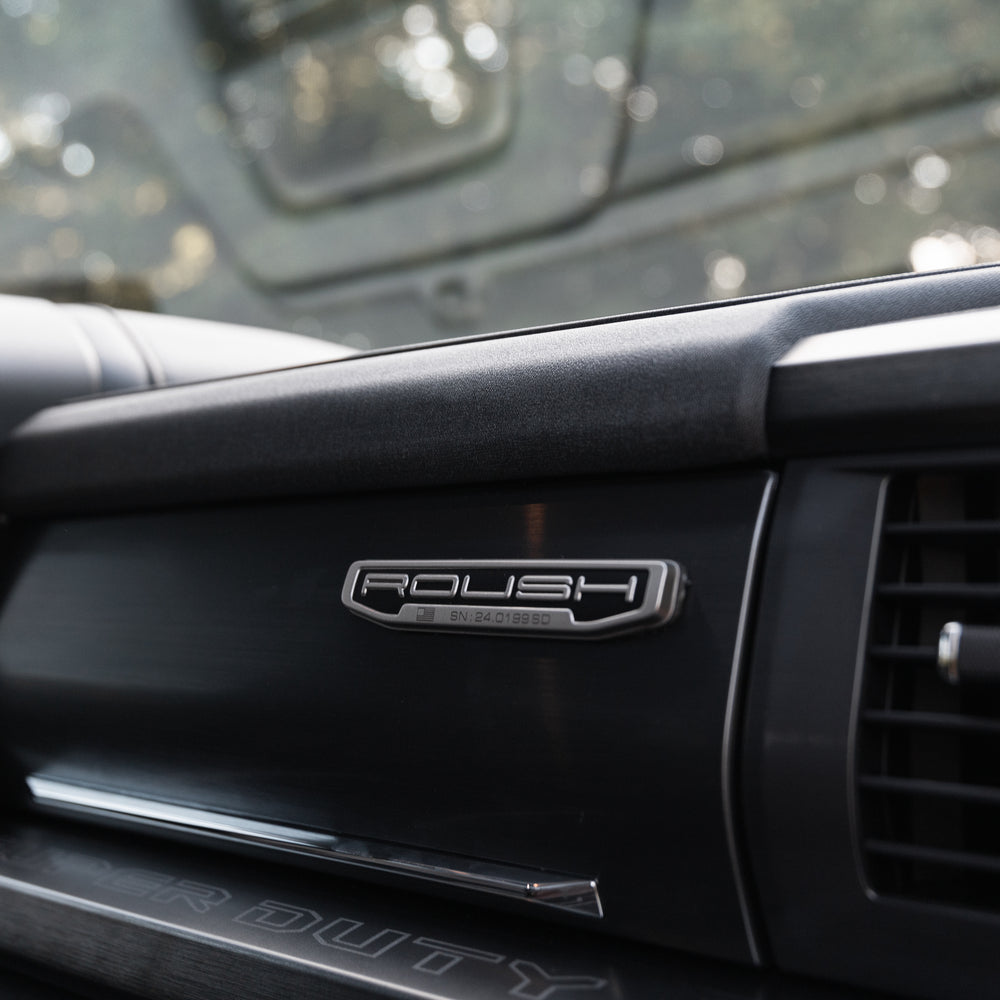 
                  
                    Close-up of a Roush Super Duty dashboard featuring a "Roush Charged" badge, air vent, and part of the instrument panel. Trees are visible through the windshield, highlighting the truck's performance suspension upgrades.
                  
                