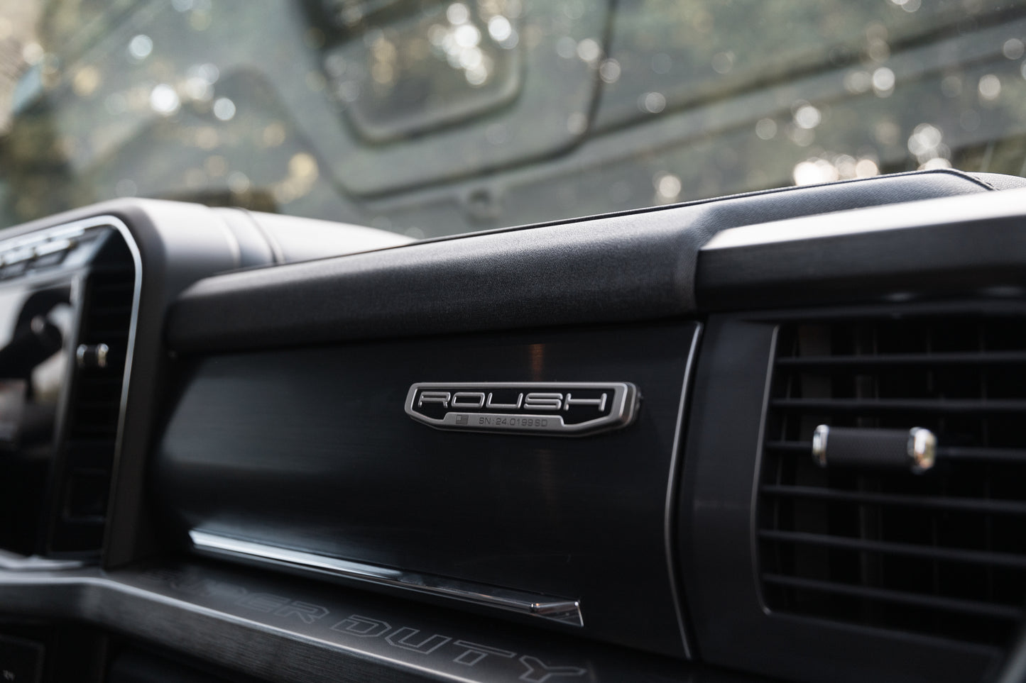 
                  
                    Close-up of a Roush Super Duty dashboard featuring a "Roush Charged" badge, air vent, and part of the instrument panel. Trees are visible through the windshield, highlighting the truck's performance suspension upgrades.
                  
                