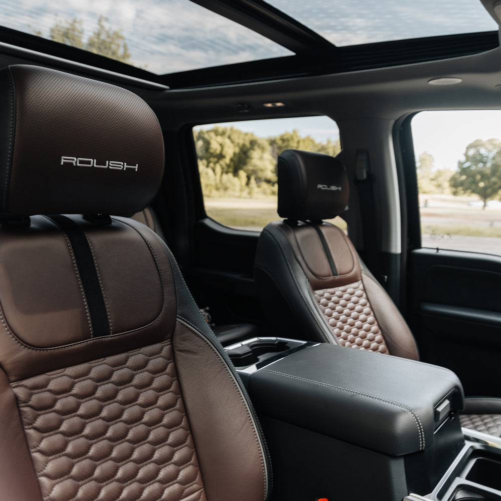 
                  
                    The interior of this Roush Super Duty boasts two brown leather seats with "ROUSH" branding, a center console, and a panoramic sunroof. Trees and sky are visible through the windows, reflecting the truck’s premium style and performance suspension.
                  
                