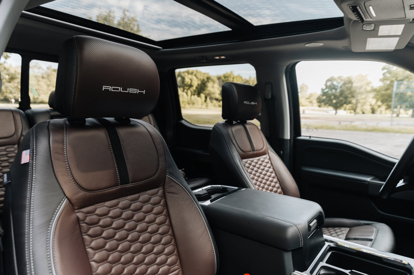 
                  
                    The interior of this Roush Super Duty boasts two brown leather seats with "ROUSH" branding, a center console, and a panoramic sunroof. Trees and sky are visible through the windows, reflecting the truck’s premium style and performance suspension.
                  
                