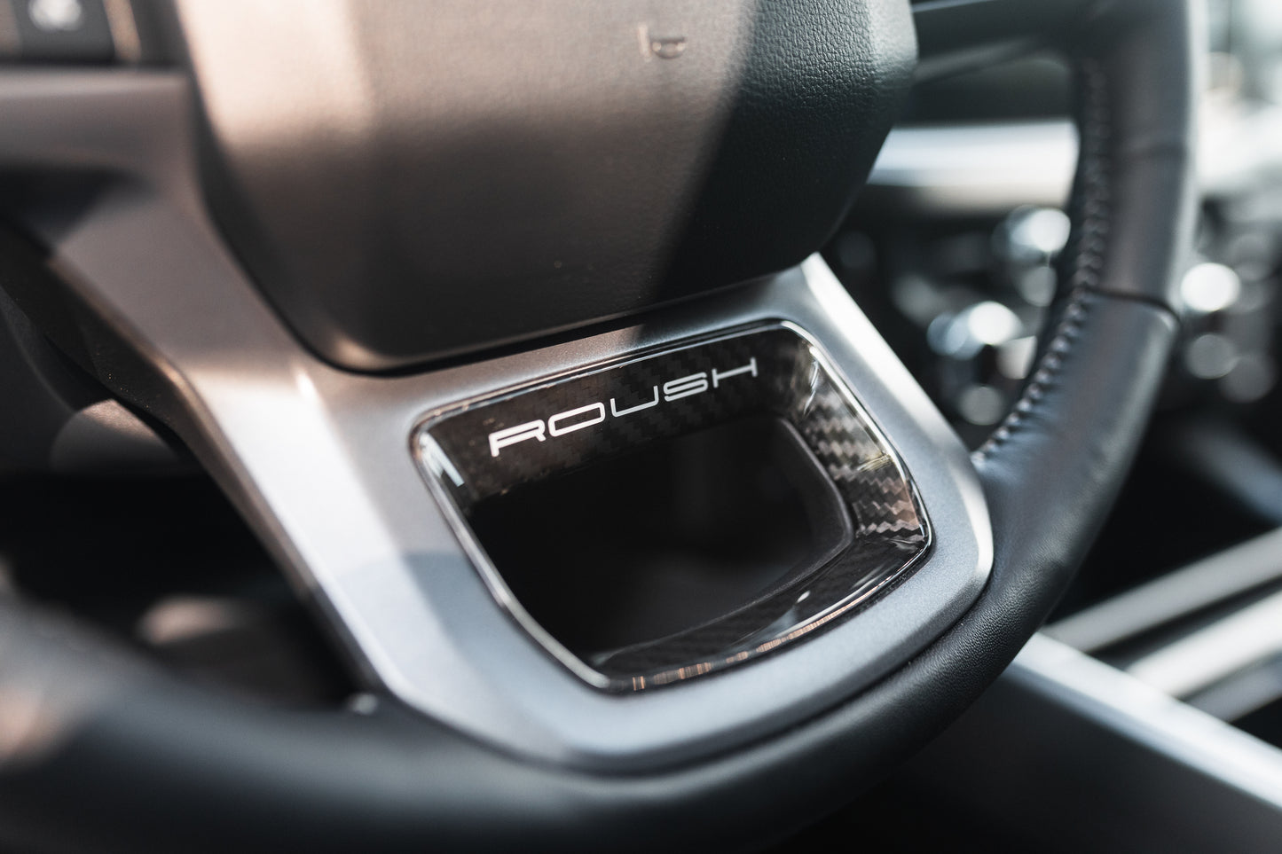 
                  
                    Close-up of a car steering wheel featuring the "ROUSH" logo on a metallic accent below the central hub, hinting at the impressive Roush Super Duty’s performance suspension.
                  
                