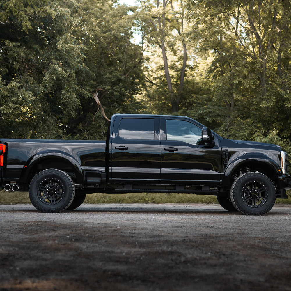 
                  
                    A black Roush Super Duty pickup truck with performance suspension is parked on a gravel surface, set against a backdrop of dense green trees and foliage.
                  
                
