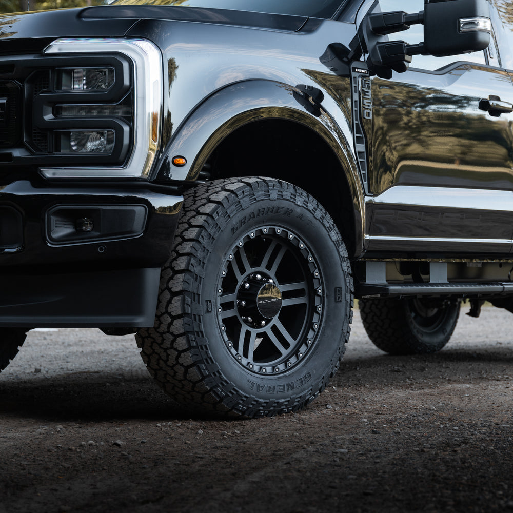 
                  
                    Close-up of the front left side of a Roush Super Duty black pickup truck parked outdoors, showcasing the tire, wheel, bumper, part of the grille, and its high-performance suspension.
                  
                