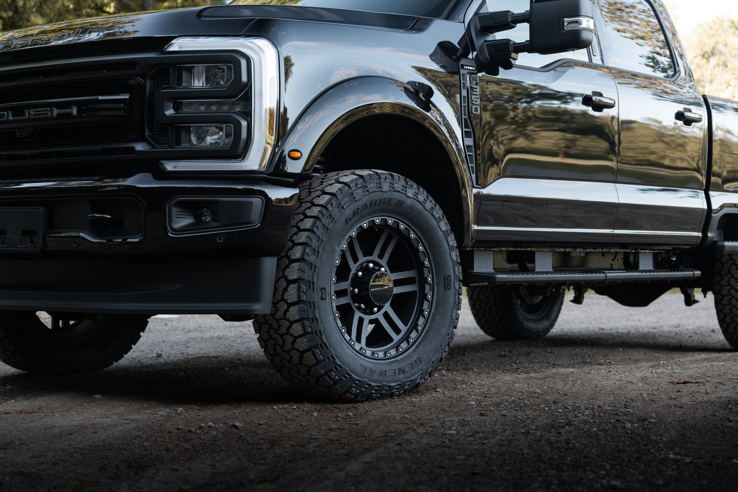 
                  
                    Close-up of the front left side of a Roush Super Duty black pickup truck parked outdoors, showcasing the tire, wheel, bumper, part of the grille, and its high-performance suspension.
                  
                
